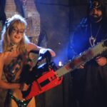 Hollywood Chainsaw Hookers 1988 Movie Scene Linnea Quigley as Samantha after The Virgin Dance of the Double Chainsaws holding a chainsaw with Gunnar Hansen as the cult leader cheering her in the background